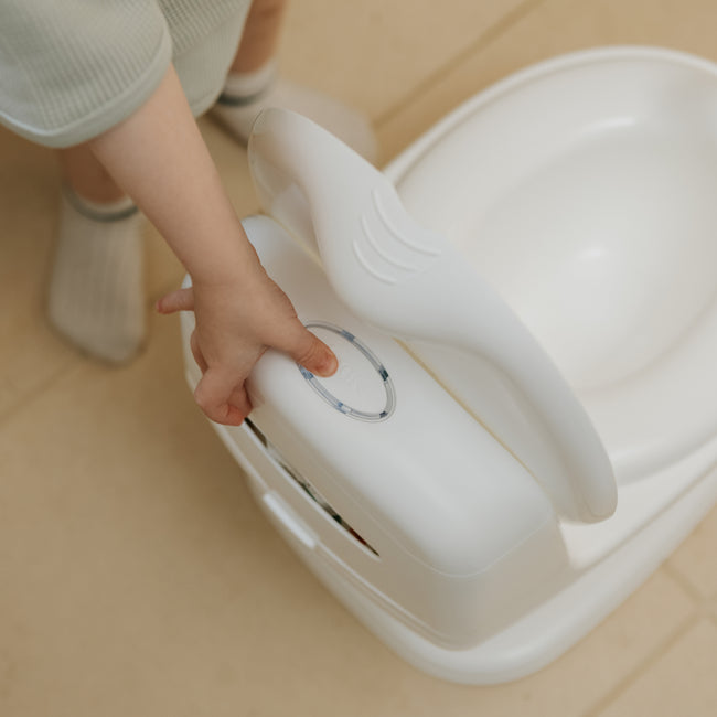 A child’s hand holds the handle of a Nuby My Real Mini Toilet Potty on a tiled floor. The child is wearing light-colored socks and pants.