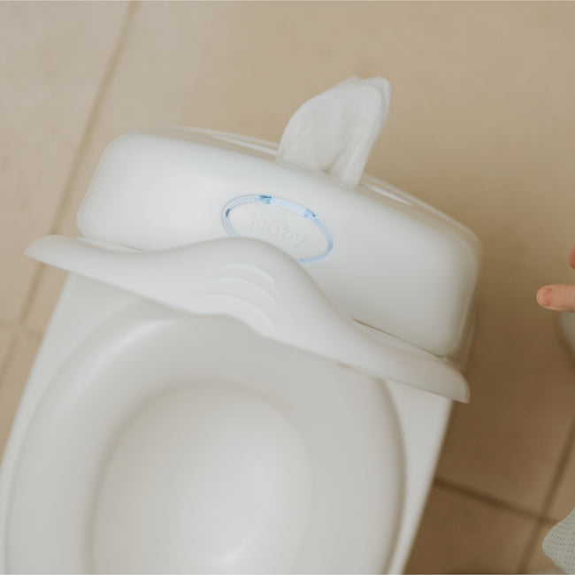 The Nuby My Real Mini Toilet Potty with a built-in wipe dispenser sits on a tiled floor. A small child’s hand is visible at the right edge of the image.