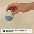A hand holds a seal-shaped thermometer from the Nuby Baby Healthcare and Grooming Kit above bathwater, its bottom half blue. The thermometer turns light blue when the water is too hot, ensuring safe bath time for your baby.