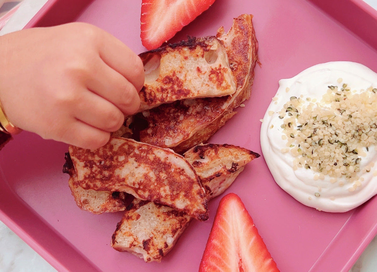 Little hands reaching for banana and strawberry French toast on a pink plate.