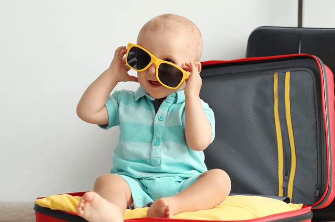 newborn essentials blog cover including a baby wearing sunglasses and being sat inside a suitcase.
