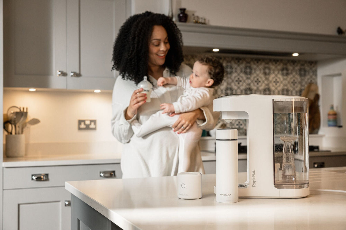 A Mother holding their baby in front of a laid out formula feeding set created by Nuby that is sat on top of a kitchen table.