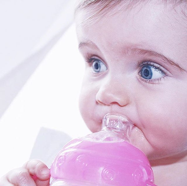 A baby drinking from a Nuby bottle that has a pink top. They are looking away from the camera while drinking.