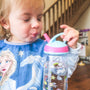 A  baby taking a sip from a cup while stood in front of a staircase. She is holding a pink and blue lidded bottle.