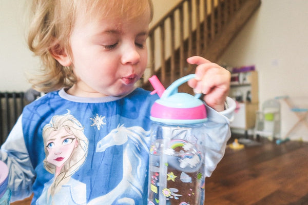 A  baby taking a sip from a cup while stood in front of a staircase. She is holding a pink and blue lidded bottle.