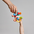 An adult hand passes the Twist Ball Teether to a baby’s outstretched hand against a plain light gray background.