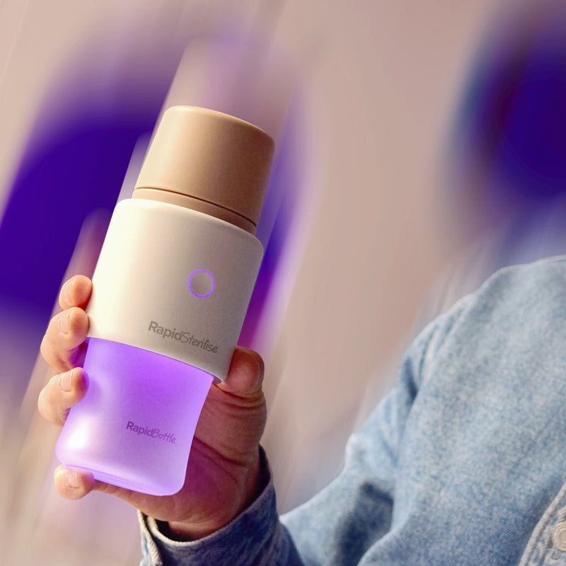 A photograph of a man's hand holding the RapidBottle while being sterilised by the Nuby RapidSterilise+. This is making the glass baby bottle look a bright violet colour. The bottle has a beige lid.