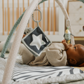 A baby lies on the Nuby UK Animal Adventures Nursery Bundle play mat, gazing up and reaching for a hanging star-shaped toy beneath gentle nursery lighting.