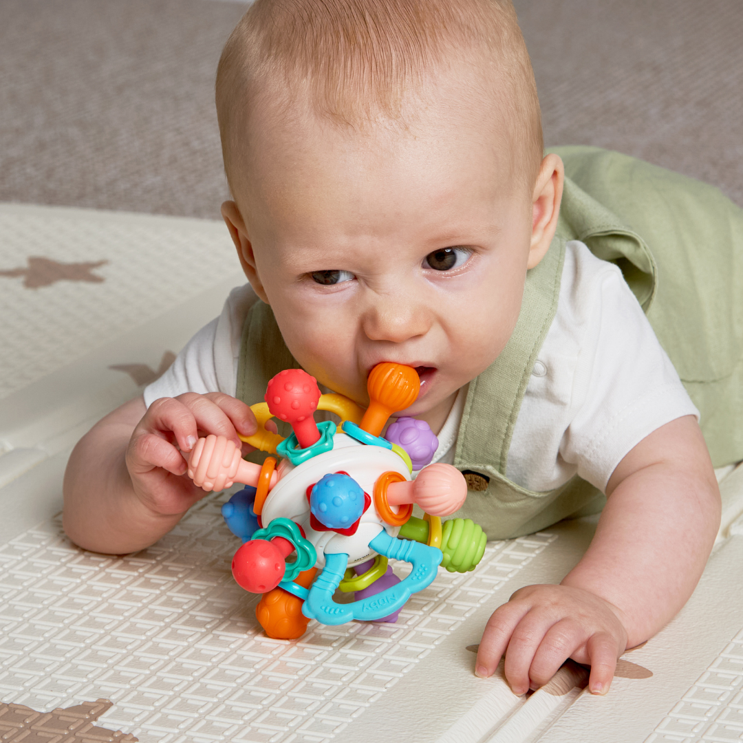 A baby lying on a play mat grasps and gnaws on the Twist Ball Teether, which features an array of shapes and textures, providing a soothing and engaging teething toy for sore gums.