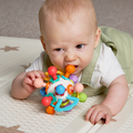 A baby lying on a play mat grasps and gnaws on the Twist Ball Teether, which features an array of shapes and textures, providing a soothing and engaging teething toy for sore gums.