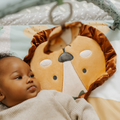 A baby enjoys the cozy Animal Adventures Nursery Bundle by Nuby UK, lying beside a plush lion face and gazing up at a hanging toy. Soft colors and gentle light create a playful, comforting scene.