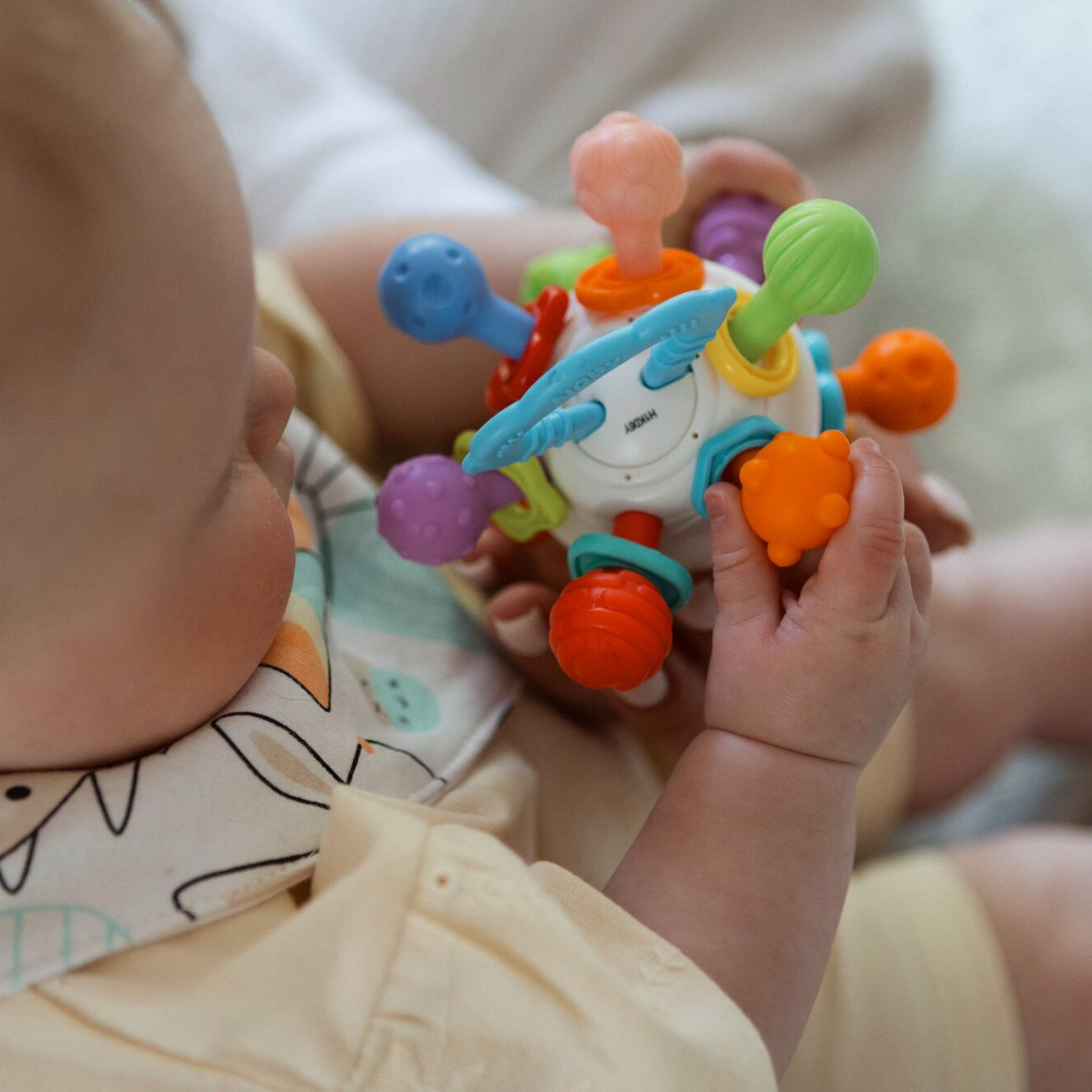 A baby sits wearing a bib and light-colored clothing while holding and exploring the colorful, multi-textured Twist Ball Teether.