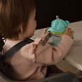 A young child in a high chair sits at a table, wearing a light pink sweater with ruffled sleeves, and holds the Nuby UK Character Sippy Cup.