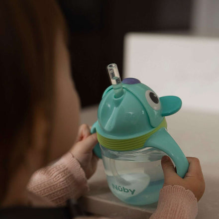 A young child holds a turquoise Nuby UK Character Sippy Cup with a weighted straw, shaped like an animal, at a light-colored table. The no-spill toddler cup displays the Nuby UK brand name.
