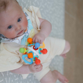 A baby in a yellow outfit sits on a light-patterned surface, holding the Twist Ball Teether—a colorful toy with various shapes and textures—and looks up toward the camera.