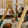 A baby relaxes on the Nuby UK Animal Adventures Nursery Bundle play mat, reaching for soft animal toys hanging from the wooden gym as an adult adjusts a toy. A crib appears in the background.