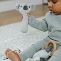 A young child enjoys the Nuby UK Animal Adventures Nursery Bundle, sitting on a patterned play mat with gym and playing with plush toys, including a soft koala rattle.