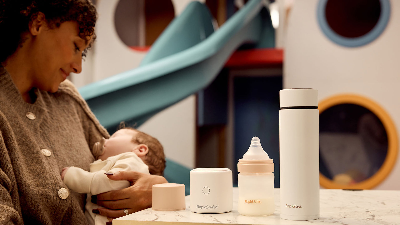 Mum holding sleeping baby, the RapidTeam on the table next to them RapidSterilise+, RapidBottle and RapidCool