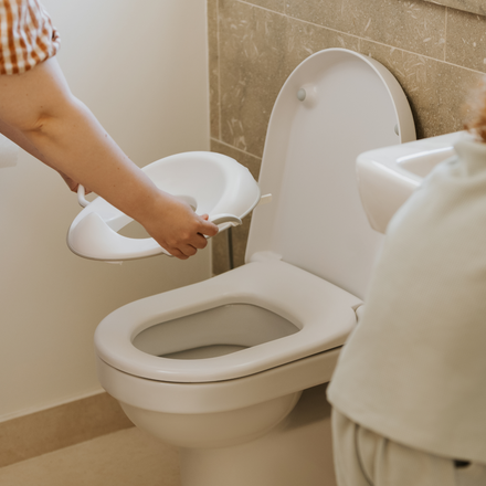 A person places a white Nuby UK Potty Training Kit Bundle toilet seat onto a standard toilet in a bathroom with beige tiles.