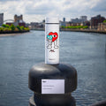 A white bottle with colorful cartoon hearts stands on a black post by a river, cityscape and cloudy sky behind. A small label reads: Nuby RapidCool x Mul portable formula maker, limited edition of 500 units.