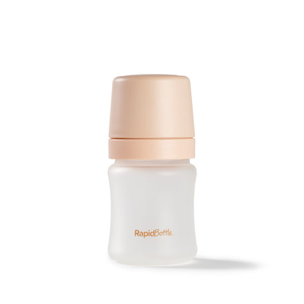 A small glass baby bottle, the RapidBottle Anti-Colic Feeding Bottle by Nuby UK, featuring a wide neck, beige cap, and orange "RapidBottle" print on the front.