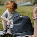 A young child with curly red hair takes a Nuby Gotta Go Travel Urinal, featuring a leak-proof design, from a gray backpack while sitting on grass outdoors. Another person is partially visible nearby on a sunny day.