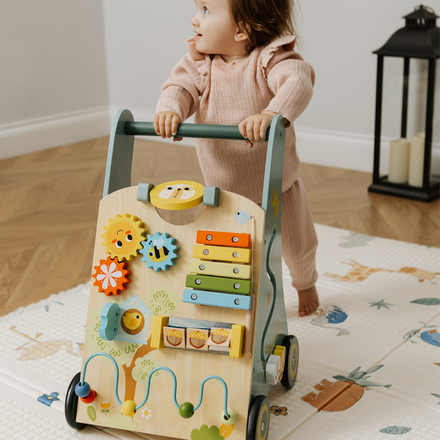 A young child in pink pajamas stands on a play mat, holding the Nuby Wooden Baby Walker, which features gears, a xylophone, and playful elements. A black lantern can be seen in the background on the wooden floor.