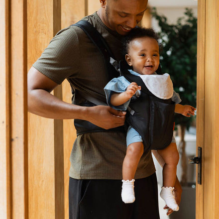 The Comfort Carrier by Nuby. A baby and their Father are interacting with the carrier in a living space with a door to the right. The Father is smiling at their baby.