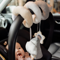 A baby lies in a car seat, gazing up and touching its mouth while the Nuby Penguin Activity Spiral Travel Pram Toy is attached to the handle for interactive fun on the go.
