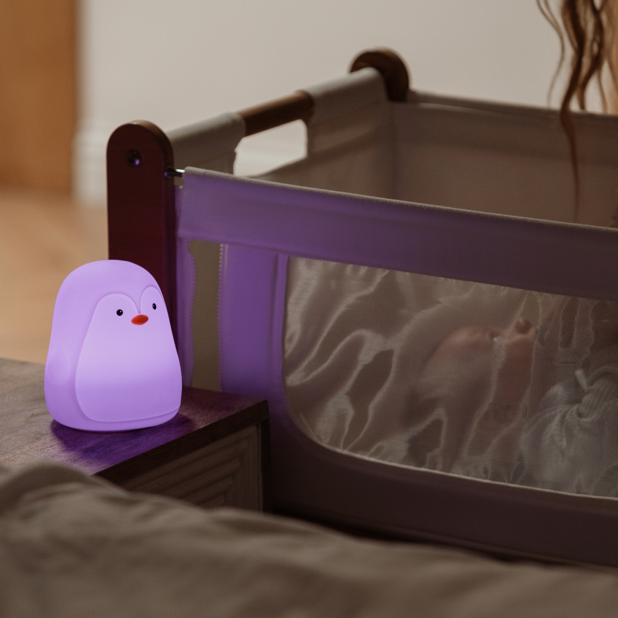 The Nuby Penguin Colour Changing Night Light glows softly on a bedside table next to a baby sleeping in a crib with mesh sides, creating a peaceful, comforting bedtime scene.