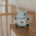 The Nuby Hippo Bath & Room Thermometer, featuring a blue hippo design and digital display reading 23°C, sits on a wooden surface indoors with a blurred wooden crib in the background.