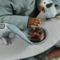 A young child in a green outfit explores the Nuby UK Animal Adventures Nursery Bundle, reaching for a small round mirror and plush animal toys on the soft, polka-dotted play mat, their hand and reflection visible in the mirror.