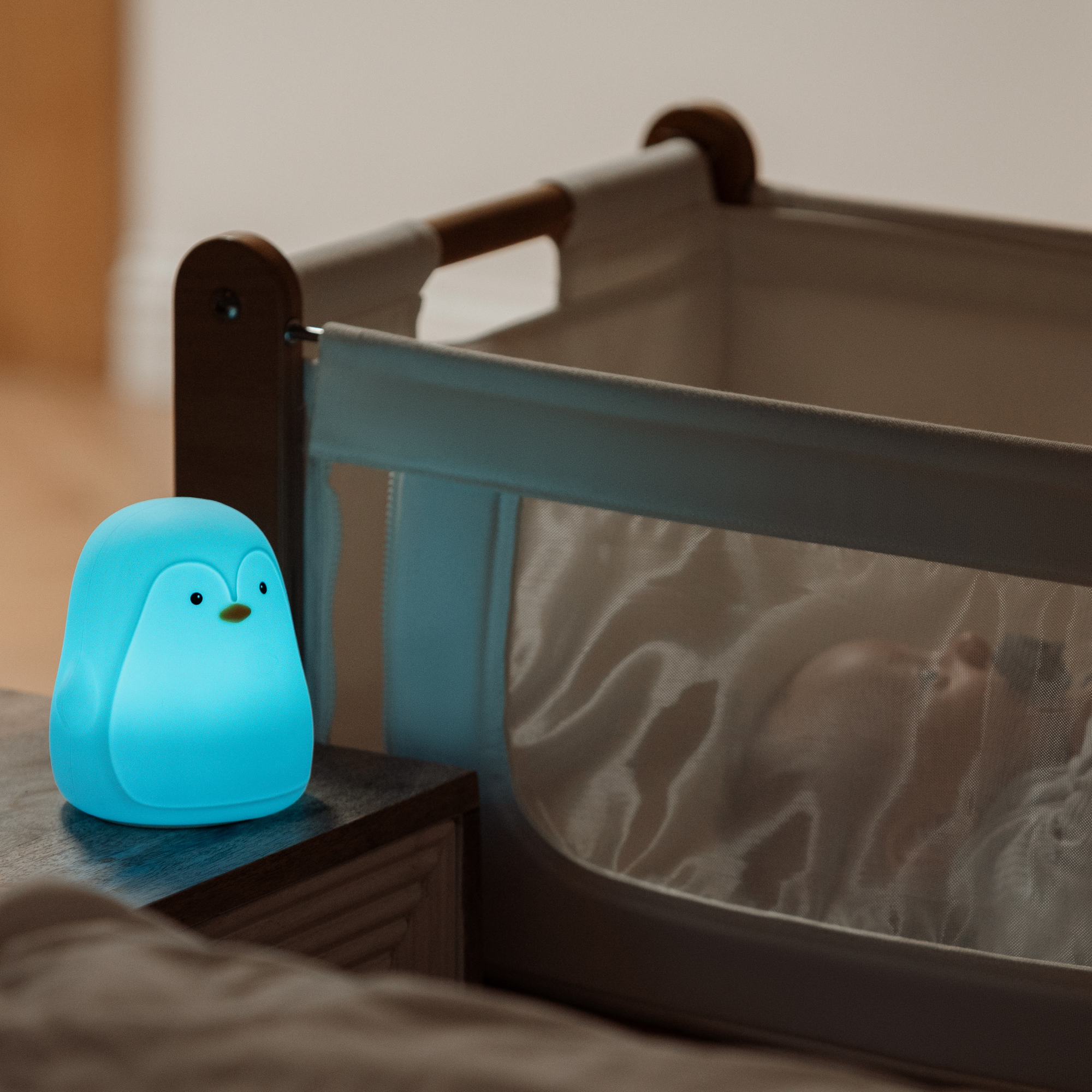 The Nuby Penguin Colour Changing Night Light sits on a table beside a sleeping baby, filling the room with a calming glow from its gentle silicone design.