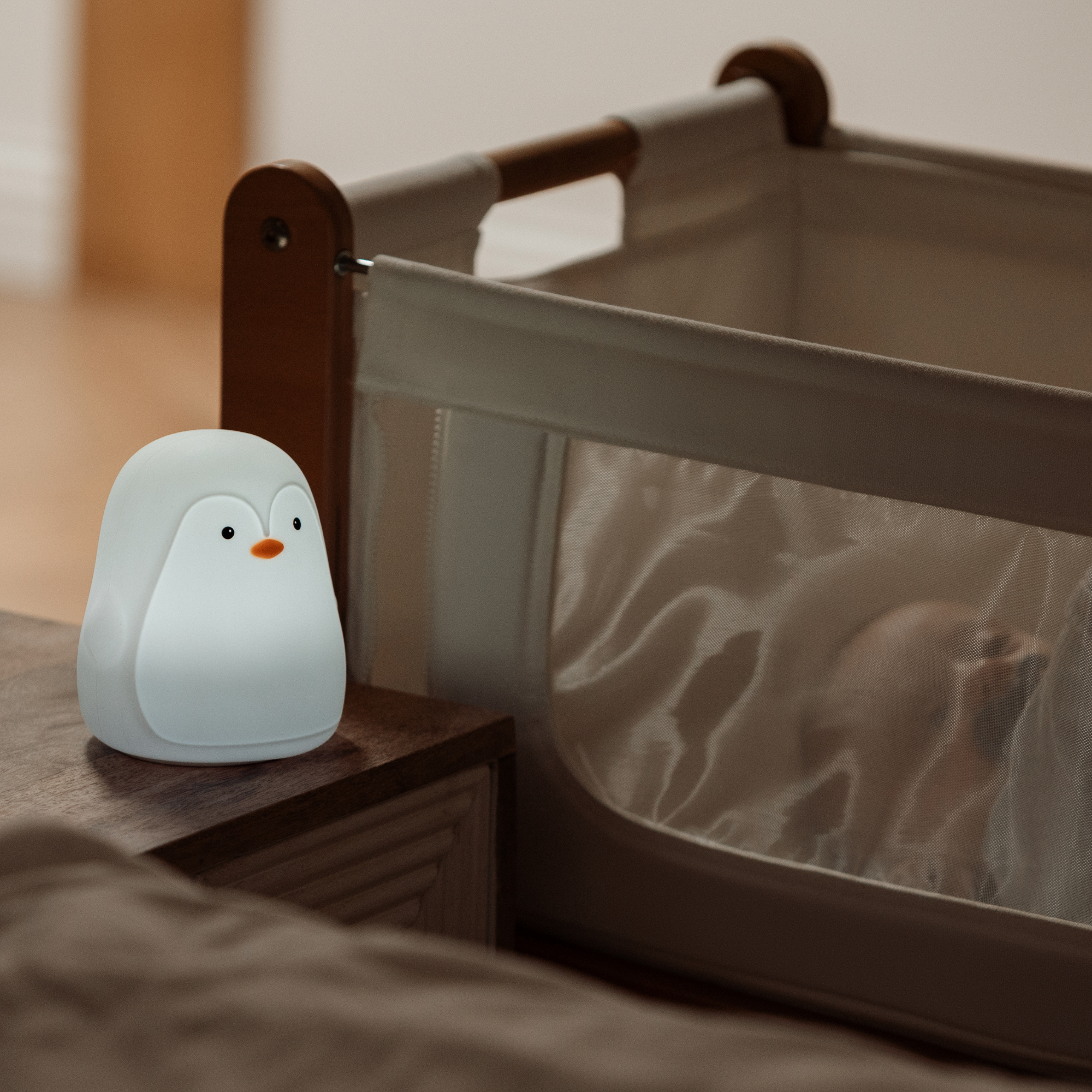 A Nuby Penguin Colour Changing Night Light glows softly on a bedside table by a crib, its soothing colours illuminating the mesh panel and gently lighting the sleeping baby.