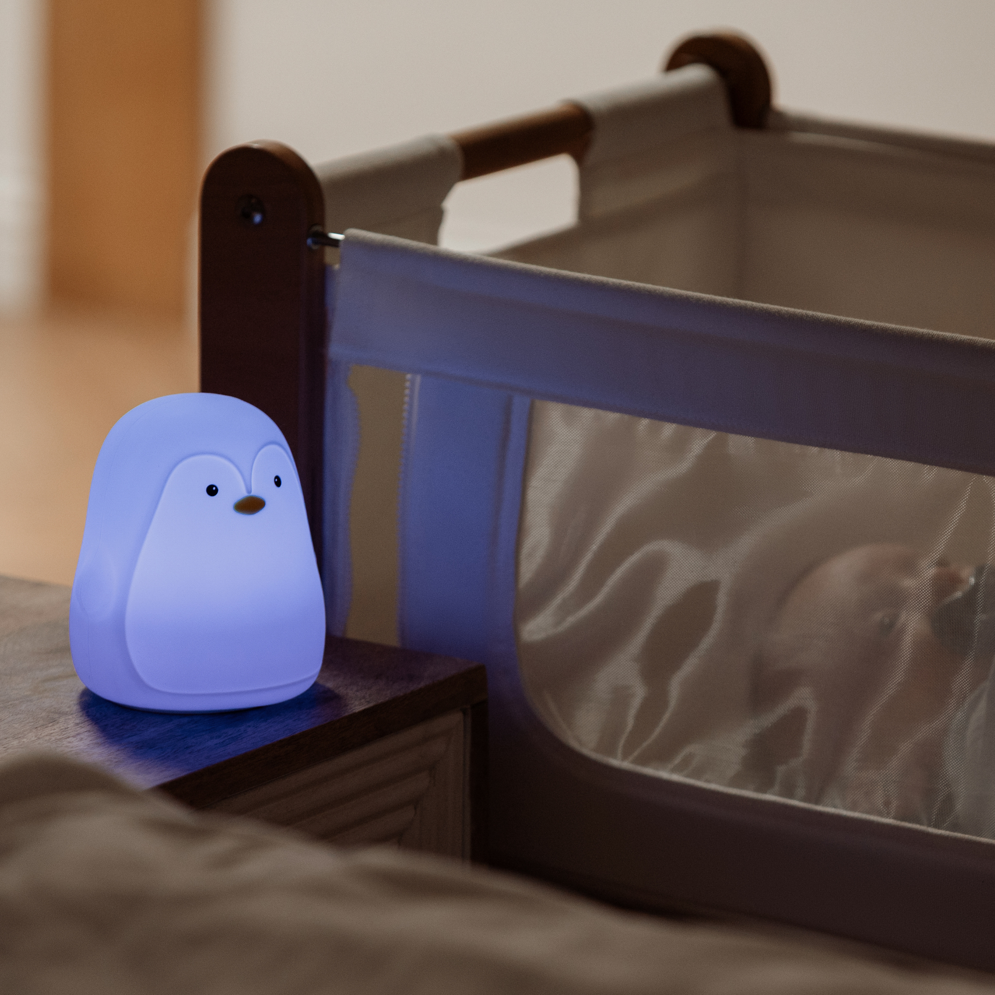 The Nuby Penguin Colour Changing Night Light, a soft blue penguin-shaped lamp, glows on a bedside table, gently illuminating a sleeping baby with soothing colours.