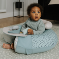 A young child with curly hair sits on the floor in a blue-patterned Nuby UK Animal Adventures Nursery Bundle seat, looking up with a surprised expression, holding soft toys and wearing a green long-sleeve outfit.