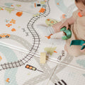 A baby, wearing socks, sits on the Nuby Foldable Play Mat, Road Map edition—decorated with train tracks and cars—holding a toy, while only their lower half is visible and toy blocks are scattered nearby.