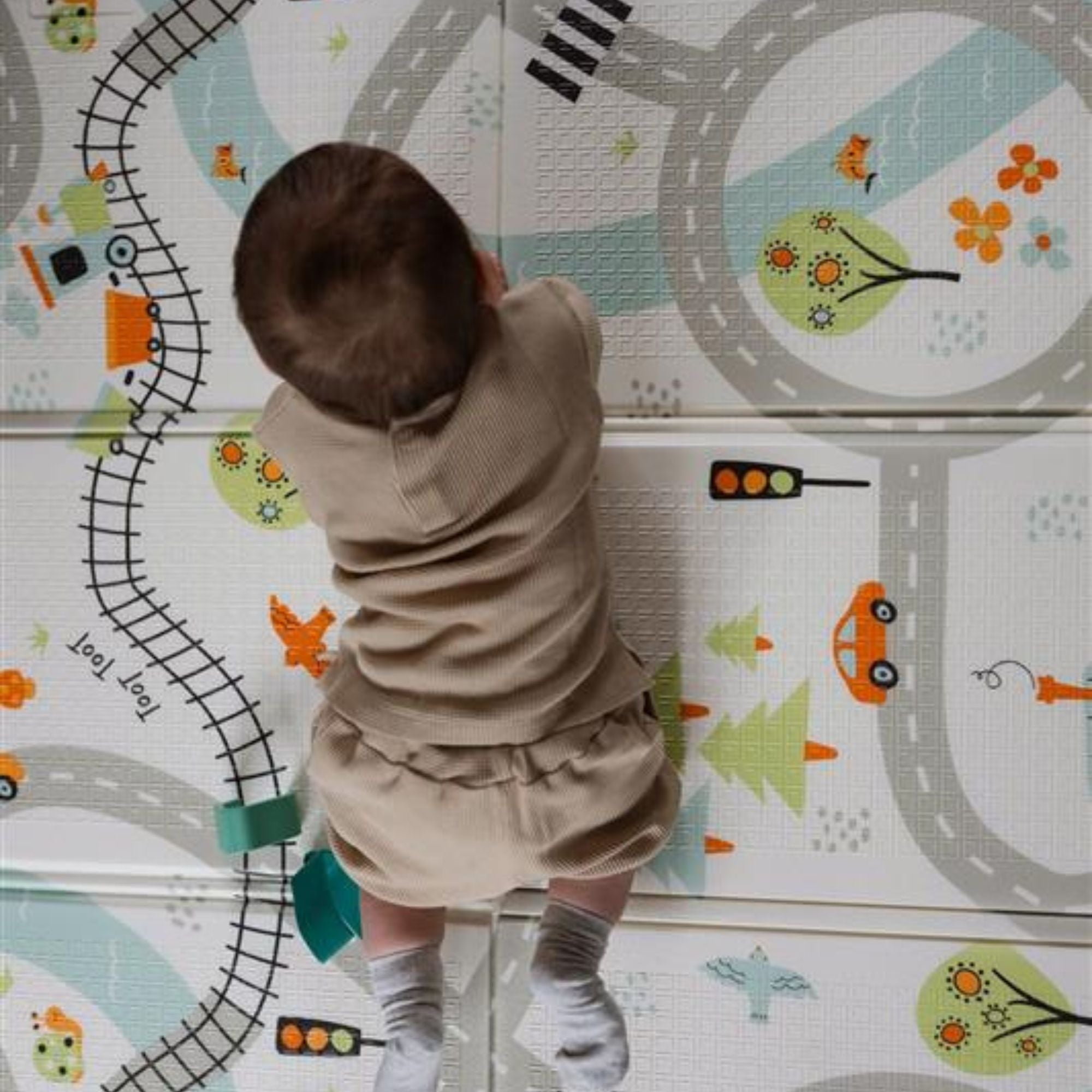 A baby in beige clothes and socks crawls on the Nuby Foldable Play Mat, Road Map, featuring reversible soft designs with colorful roads, train tracks, cars, trees, birds, and traffic lights.
