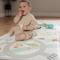 A baby in beige sits on a Nuby Foldable Play Mat, Road Map, holding cheese. Toys and play kitchen items are scattered nearby on the floor.