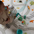 A baby in a beige outfit and white socks plays with colorful cups on the Nuby Foldable Play Mat, Road Map, which features roads, train tracks, cars, trees, and tents.