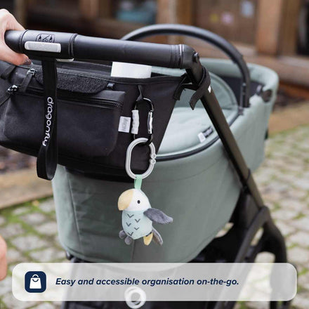 A hand pushes a stroller with the Nuby Pram Organiser Bag and a toy bird attached, on a cobblestone path. Text overlay: Easy and accessible organisation on-the-go.