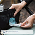 A hand pulls a baby wipe from the Nuby UK Pushchair Essentials Kit pouch attached to a stroller handle, with other pushchair essentials nearby. The pouch’s easy-access opening makes wipes quick to grab. Text: “Easy access wipes pouch.”.