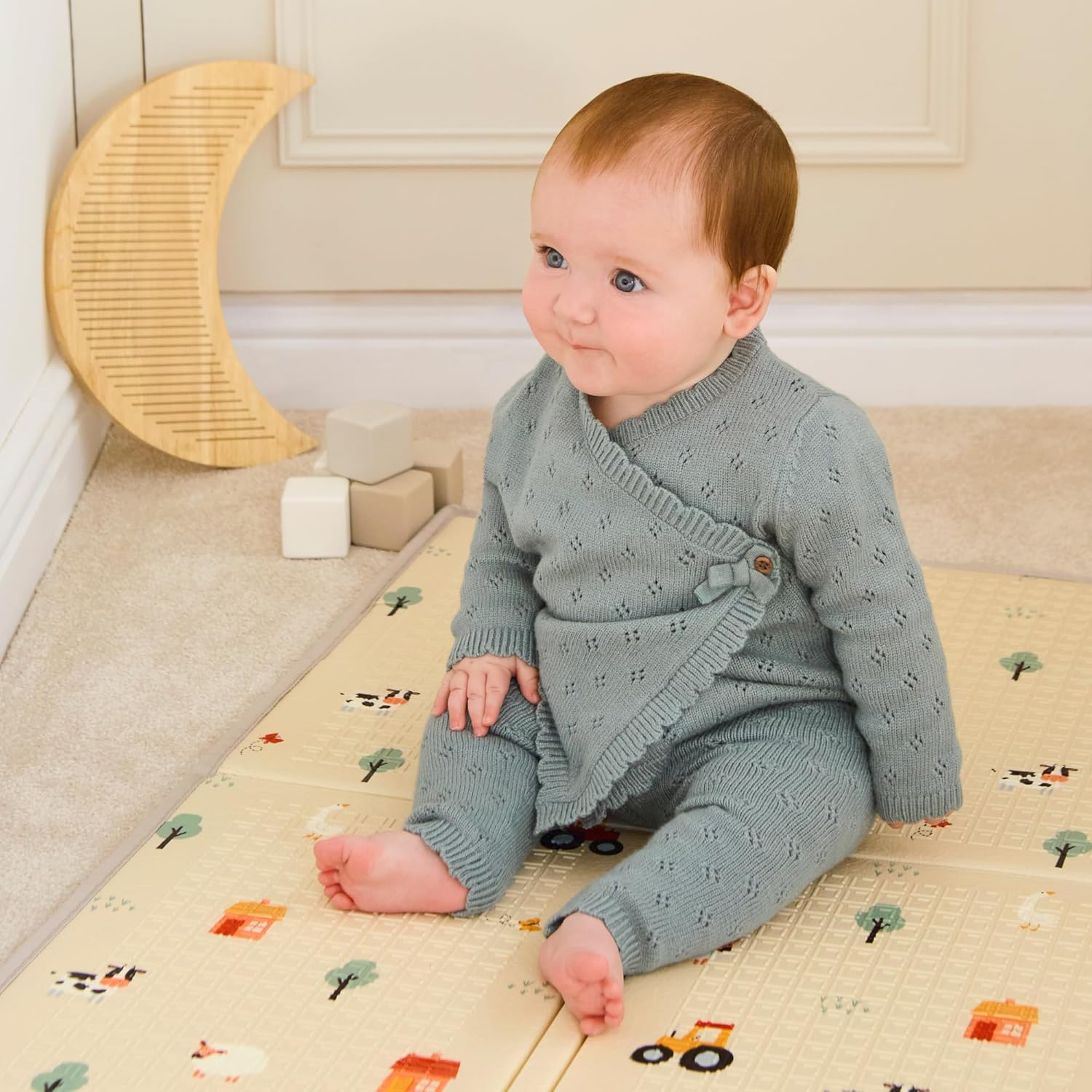 A baby with reddish-brown hair sits on a Fold N Go Play Mat featuring small trees and vehicles, dressed in a light blue knitted outfit. Wooden blocks and a crescent moon decoration are visible in the background.
