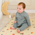 A baby with reddish-brown hair sits on a Fold N Go Play Mat featuring small trees and vehicles, dressed in a light blue knitted outfit. Wooden blocks and a crescent moon decoration are visible in the background.