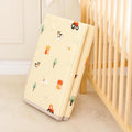 The Fold N Go Play Mat, a cream-colored cushioned foam mat with farm-themed illustrations, is folded and leaning against a wooden crib in a softly lit nursery.