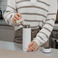 Wearing a striped sweater, someone pours cream into a white insulated travel mug on the kitchen counter near the Nuby UK RapidCool Bottle Maker and Portable UV Steriliser Bundle, with the mug’s lid placed beside it.