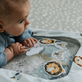 A baby in a blue sweater enjoys tummy time on the Nuby UK Animal Adventures Nursery Bundle, a vibrant play mat decorated with animal illustrations like a lion and elephant.