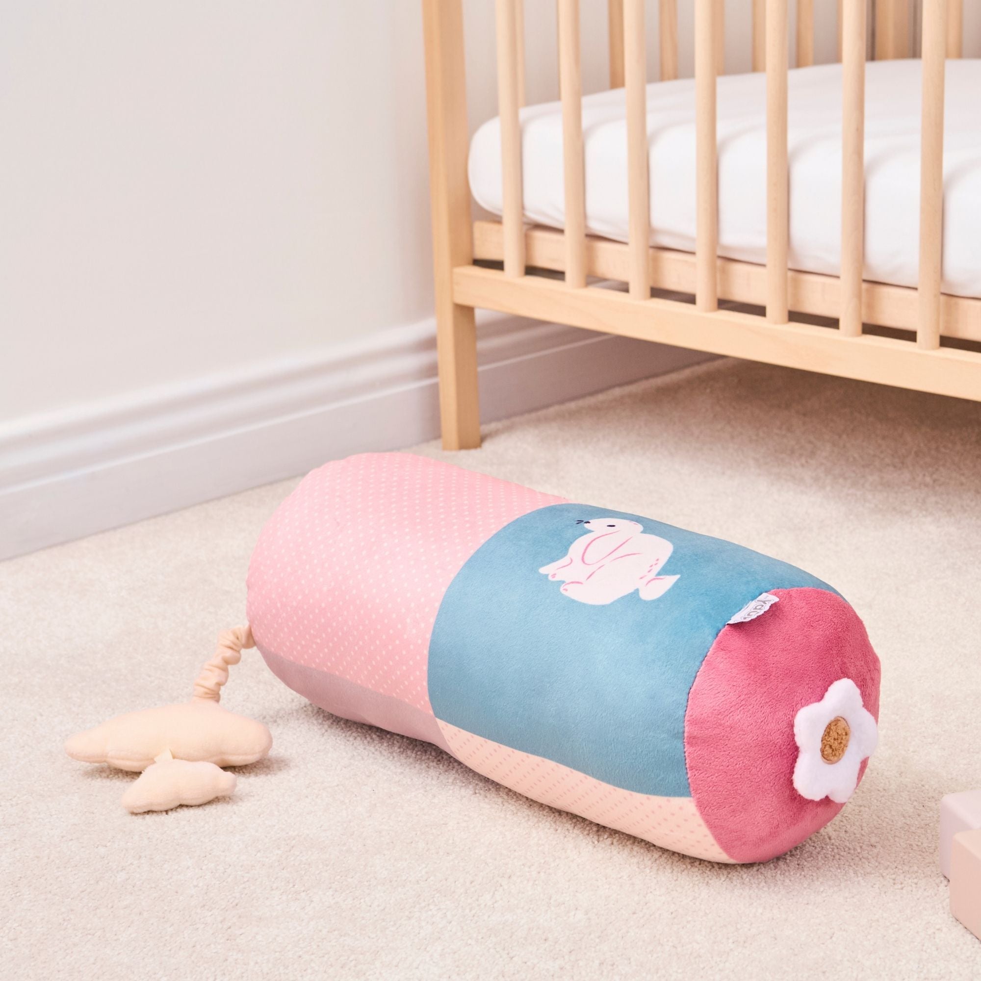 The Little Hops & Raindrops Tummy Pillow is a soft cylindrical bolster with pastel pink and blue panels, a white flower on one end, and a duck illustration on the side, pictured on the floor next to a wooden crib in a nursery.