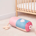 The Little Hops & Raindrops Tummy Pillow is a soft cylindrical bolster with pastel pink and blue panels, a white flower on one end, and a duck illustration on the side, pictured on the floor next to a wooden crib in a nursery.