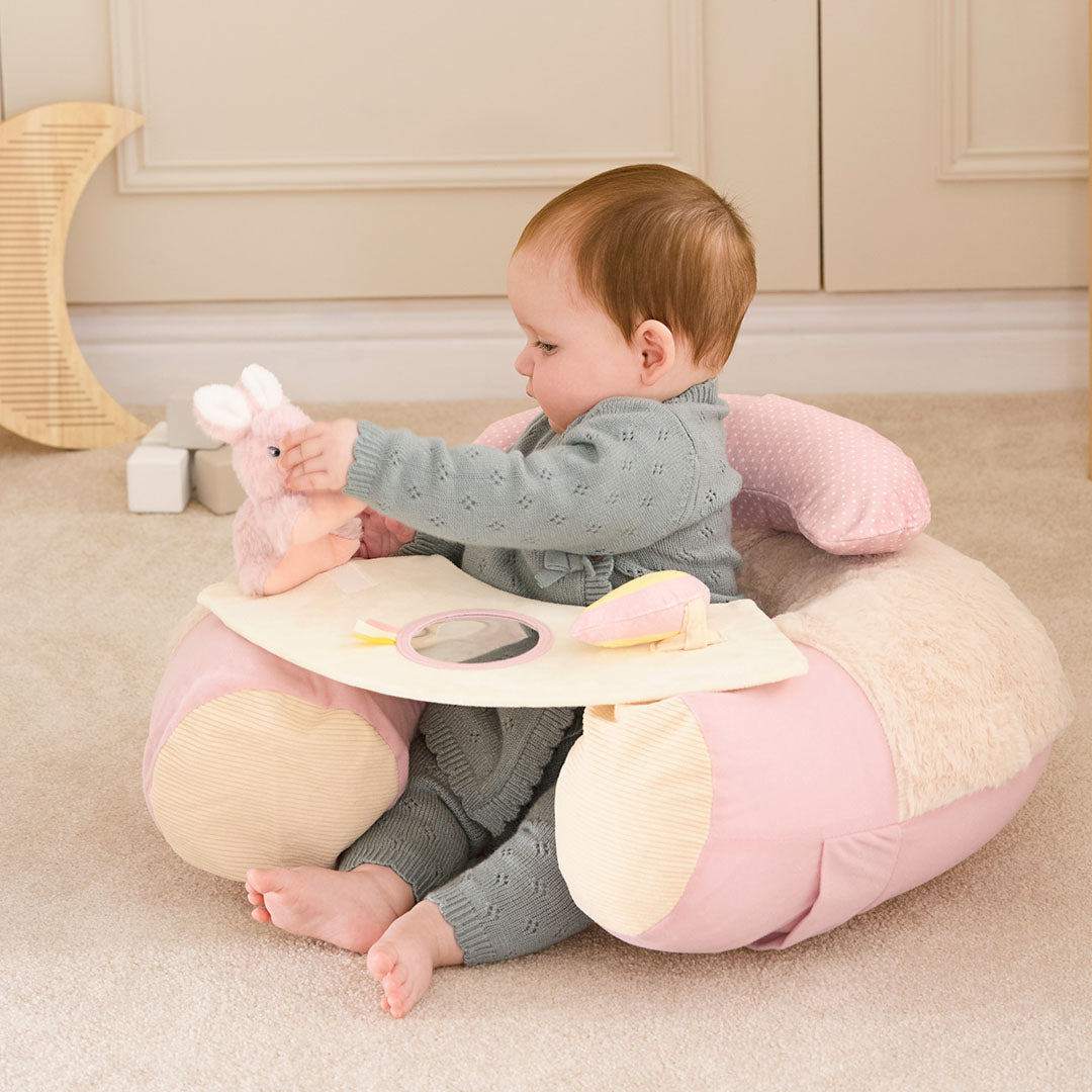 A baby enjoys cozy comfort in the Little Hops & Raindrops Sit Up Seat—in soft pink and beige—with a plush bunny toy on the built-in tray, playing happily in an inviting indoor space.